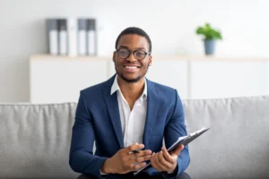 Priser 5 Portrait of Happy Black Male Psychologist Looking at Camera and Taking Notes during Therapy Session at Clinic